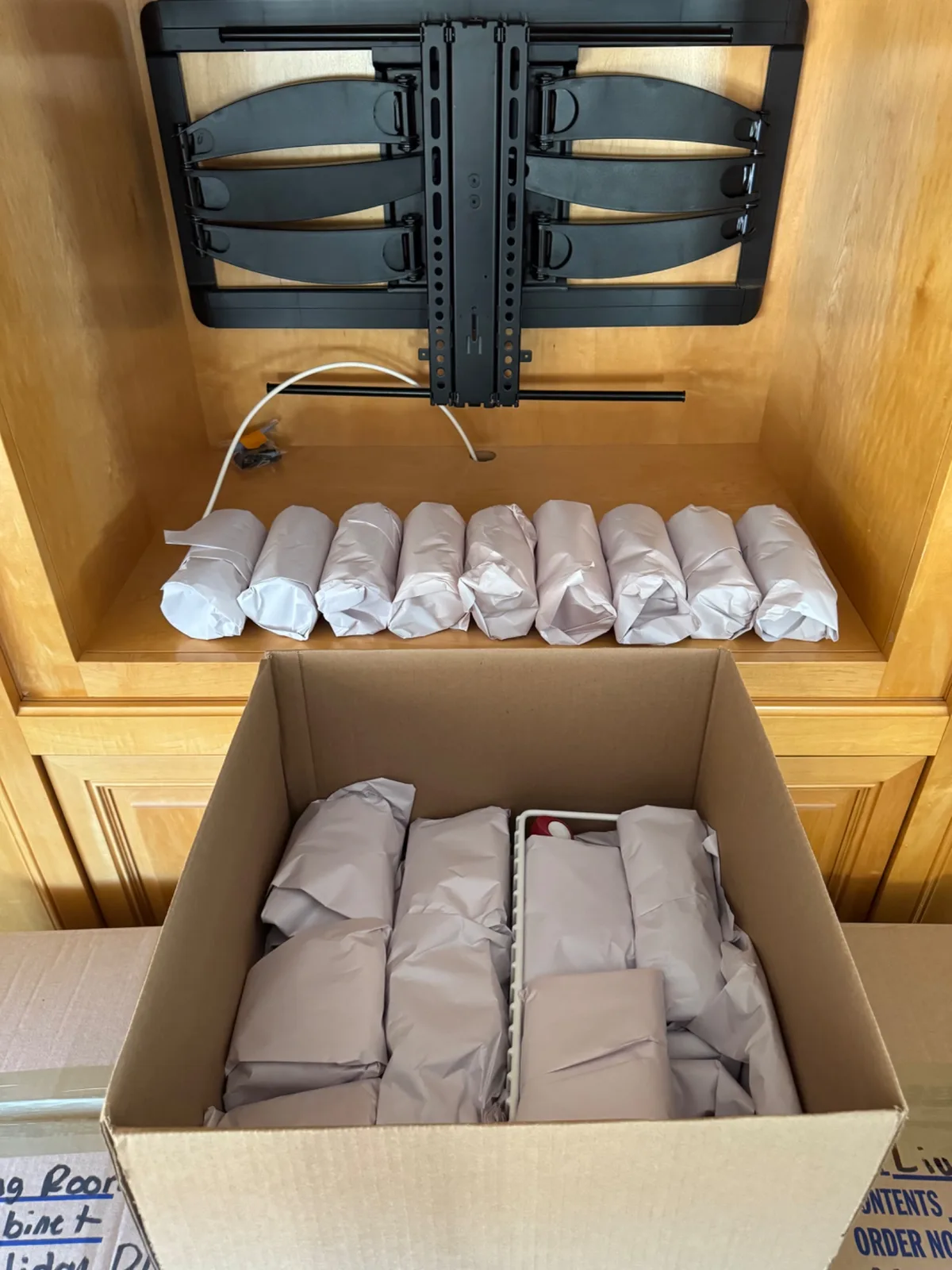 A cardboard box filled with neatly wrapped items in front of a wooden cabinet with a TV wall mount.
